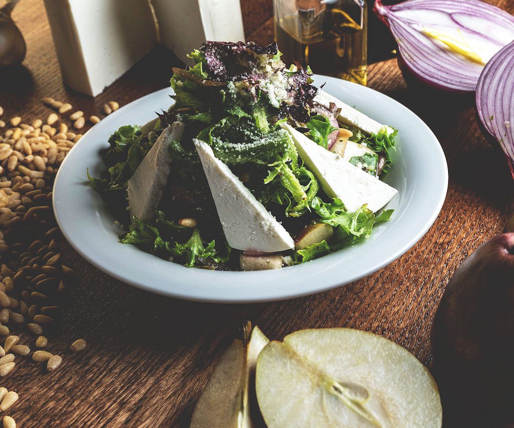 A delightful pear salad with ricotta salata, featuring slices of ripe pear arranged on a bed of mixed greens, topped with crumbled ricotta salata cheese, walnuts, and a drizzle of balsamic vinaigrette, presenting a flavorful and elegant salad option at Spumoni Gardens.