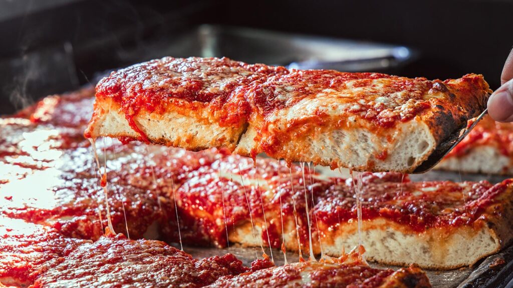 A close-up of a hand lifting a slice of thick, rectangular pizza with melted cheese and tomato sauce. Steam rises from the hot slice, with more pizza visible in the background.