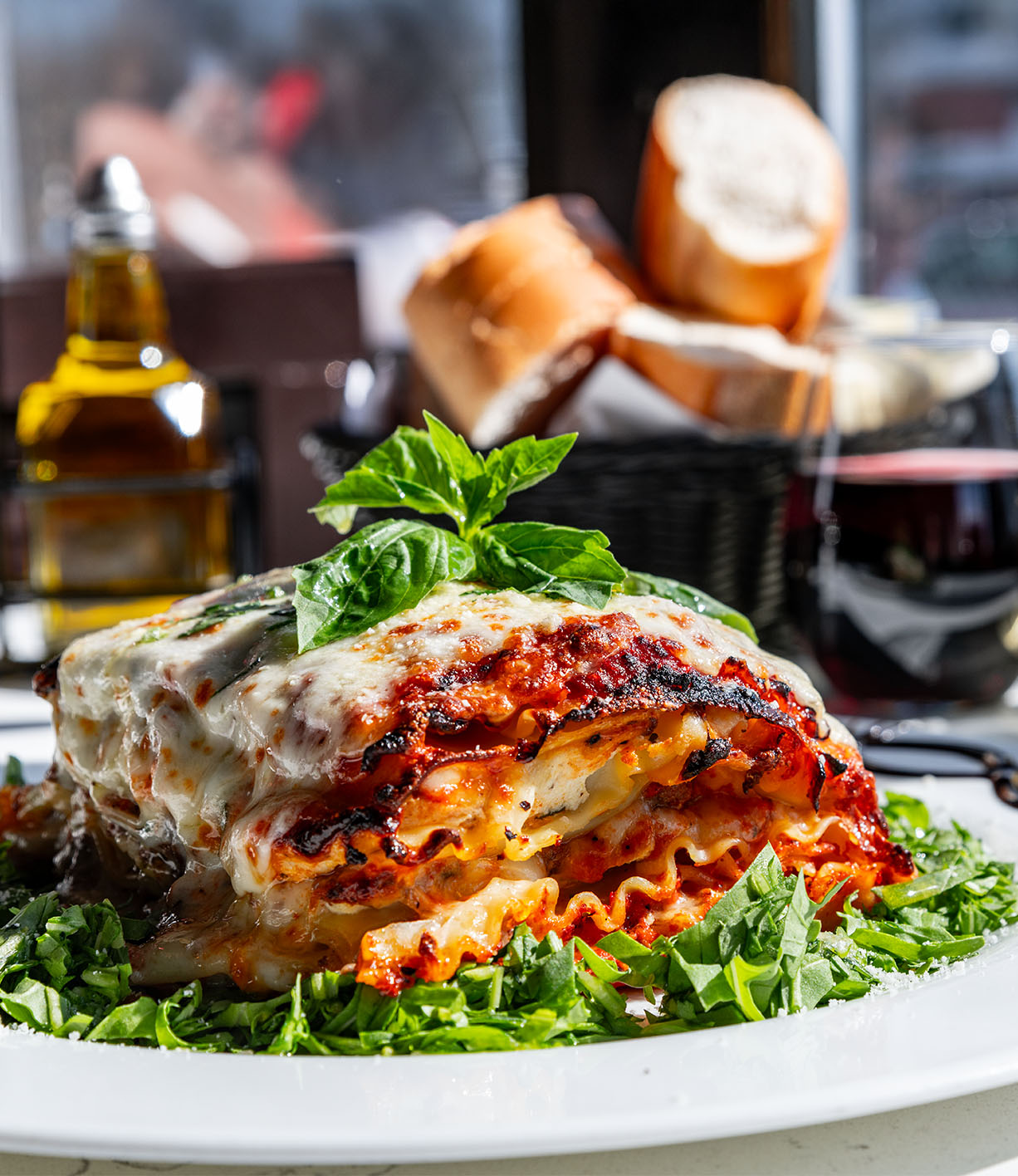 A plate of lasagna topped with basil sits on a bed of arugula. Bread and a bottle of oil are in the background.