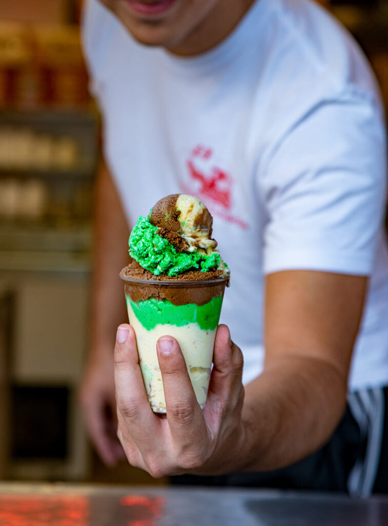 A person in a white shirt holds out a cup of layered ice cream with chocolate, green, and vanilla scoops.