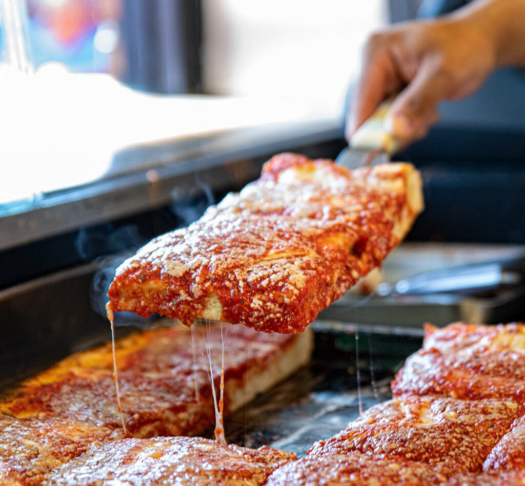 A hand holds a slice of deep-dish pizza with tomato sauce and melted cheese, showing cheese stretching as it's lifted from a pan with similar slices.