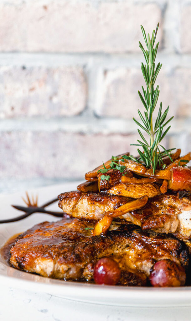 Grilled meat topped with cooked vegetables and herbs, garnished with a sprig of rosemary, on a white plate against a brick wall.