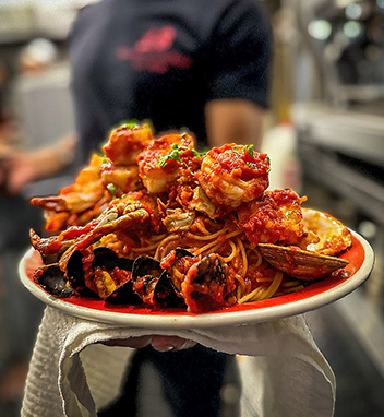 A person holds a plate of seafood pasta with shrimp, mussels, and crab in tomato sauce.