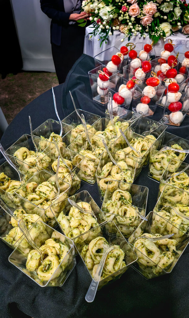 Small clear cups filled with pasta and pesto, arranged on a black tablecloth; behind them are skewers with mozzarella and cherry tomatoes, and a floral arrangement in the background.