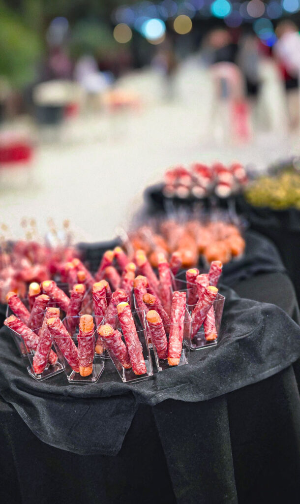 Close-up of appetizers with rolled cured meats on skewers, displayed on a table covered with a black cloth at an outdoor event.