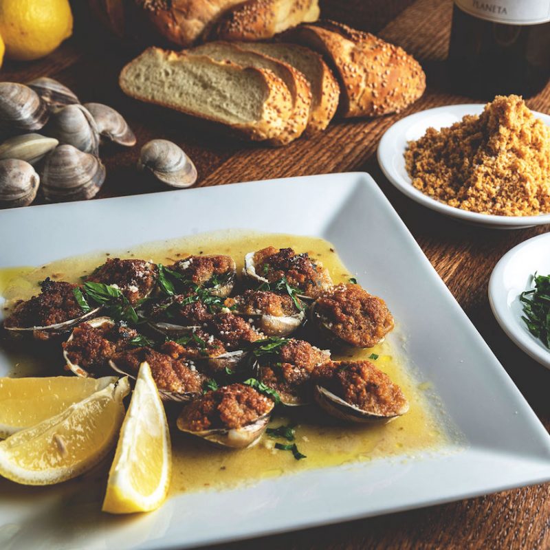 A mouthwatering plate of baked clams, showcasing golden-brown clam shells filled with a savory breadcrumb stuffing, garnished with fresh parsley, offering a delectable seafood appetizer option at Spumoni Gardens.