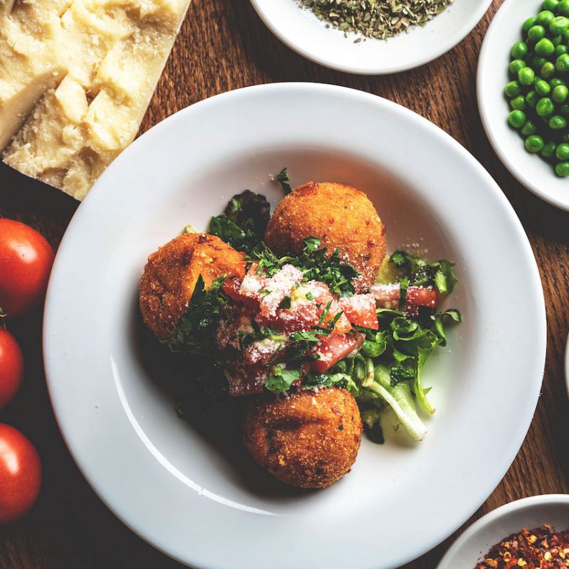 Golden brown rice balls, also known as arancini, served on a white plate surrounded by raw ingredients such as parsley, tomatoes, peas, cheese and additional garnishes, presenting a visually appealing and appetizing display at Spumoni Gardens.