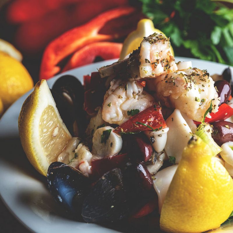A refreshing seafood salad featuring a medley of shrimp, calamari, and mussels tossed with vinaigrette dressing, presented in a white bowl, offering a flavorful and appetizing seafood option at Spumoni Gardens.